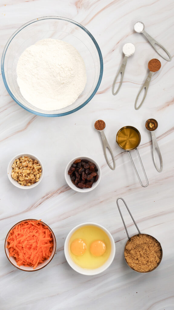 A flat lay of carrot cake muffin ingredients, including a bowl of shredded carrots, flour, eggs, sugar, spices, and oil, arranged on a wooden kitchen counter.