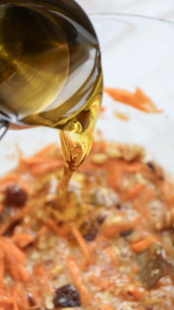 A close-up of vegetable oil being poured from a measuring cup into a mixing bowl with carrots.
