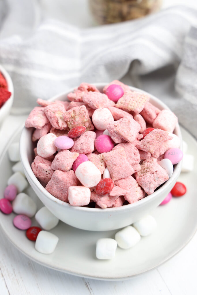 Bowl of colorful Strawberry Puppy Chow with Chex cereal, Valentine’s M&Ms, and marshmallows on a table.
