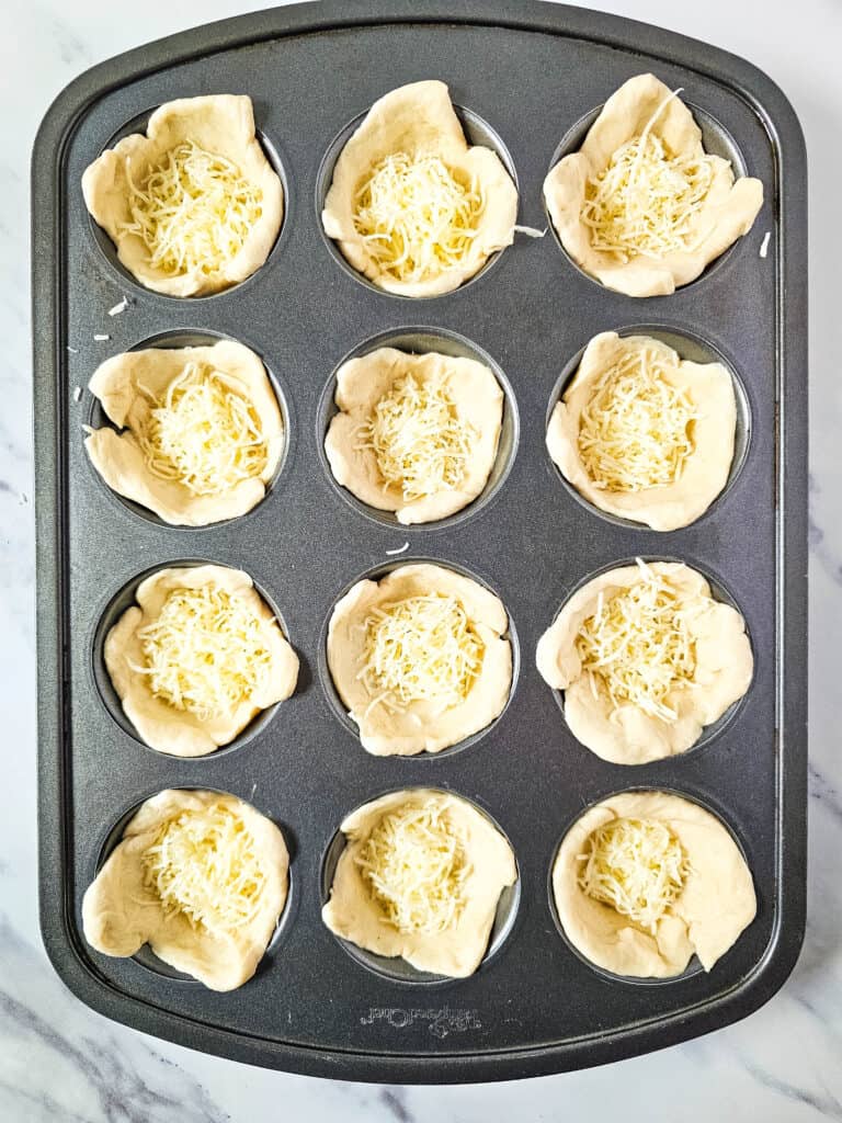 Muffin tin with dough and cheese ready for the next step in making pepperoni pizza bites.