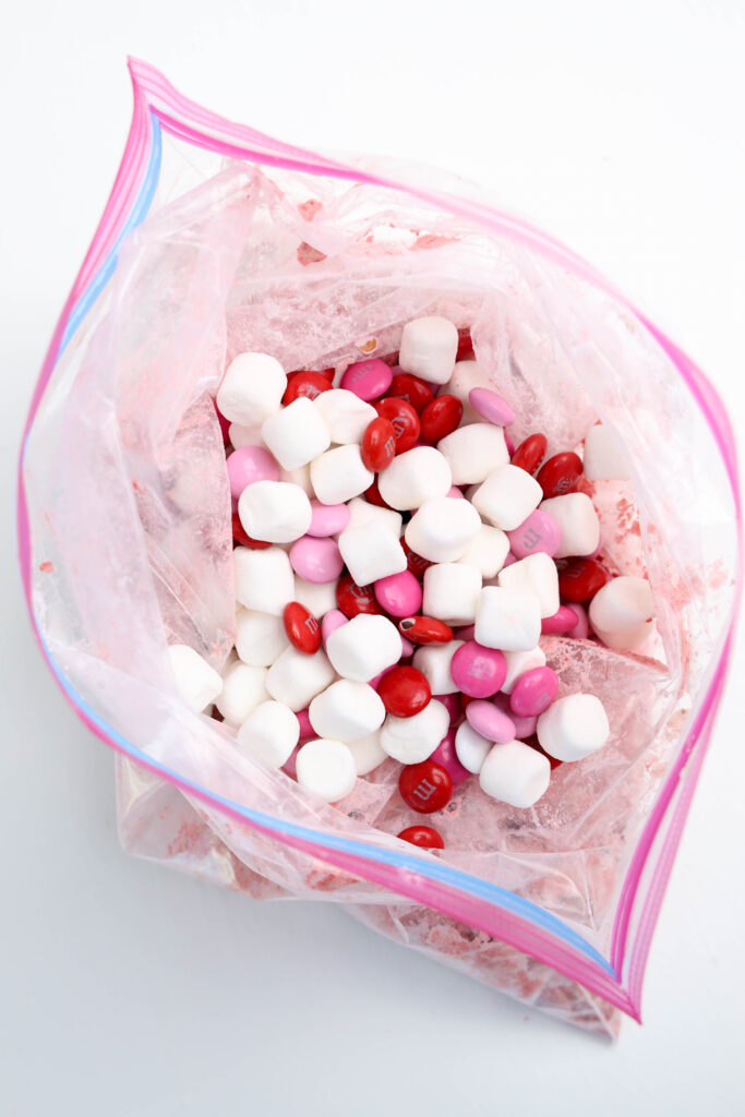 Chex cereal coated in strawberry powder being mixed with M&Ms and marshmallows.