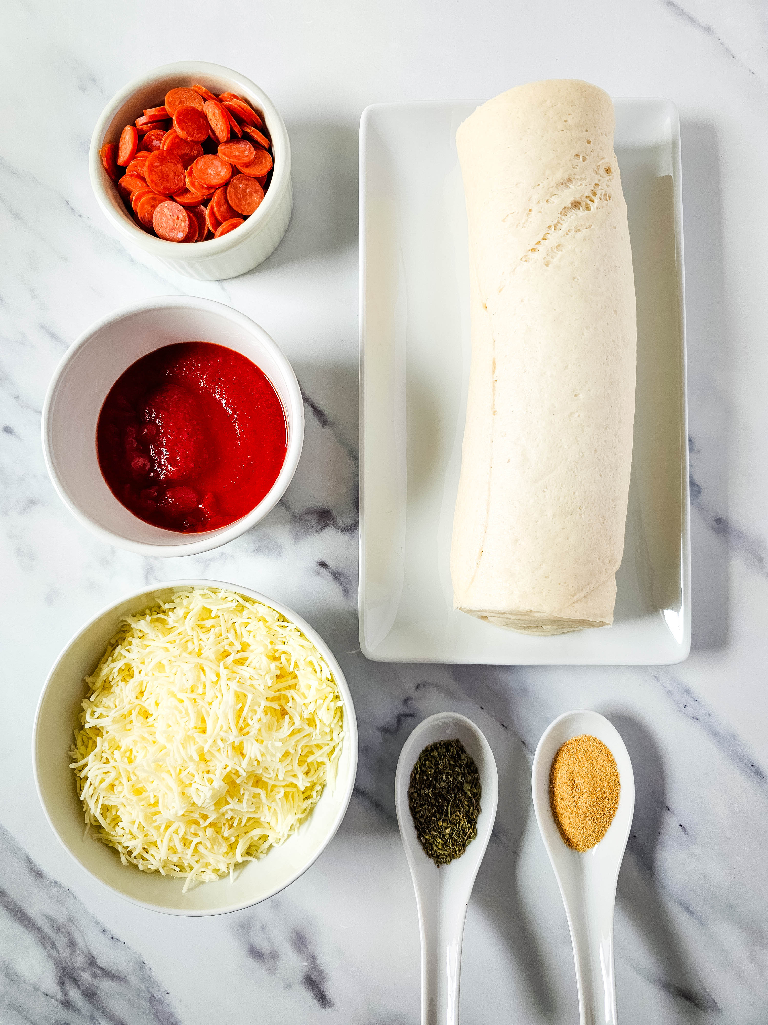 Ingredients for pepperoni pizza puffs, including pizza dough, shredded mozzarella, mini pepperoni, pizza sauce, garlic powder, and Italian seasoning, arranged on a countertop.