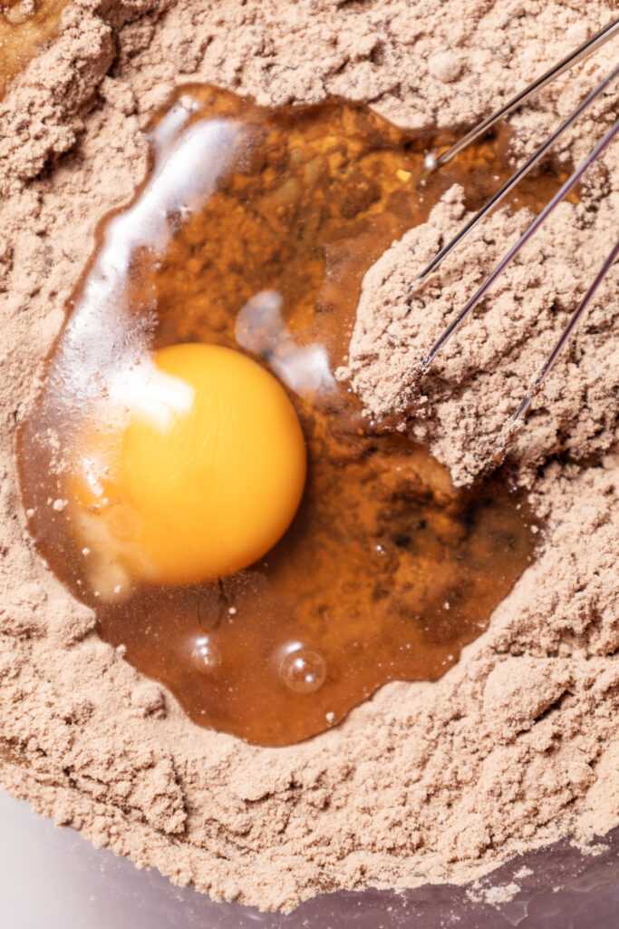 A cracked egg being added to glossy chocolate brownie batter in a mixing bowl.