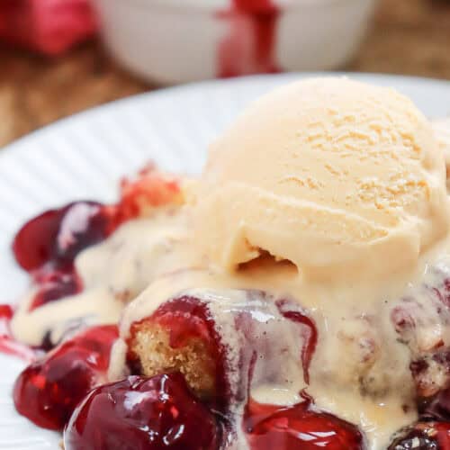 Close-up of a scoop of Cherry Dump Cake served with vanilla ice cream on a white plate.