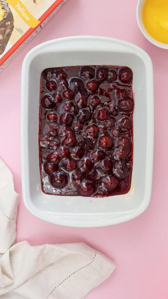 Close-up of cherry pie filling being poured into a white baking dish as the first step in making Cherry Dump Cake.